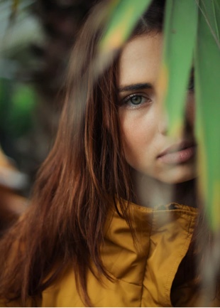 Designer, Willow, with folliage obsecuring part of her face. Looking at camera, wearing a yellow raincoat